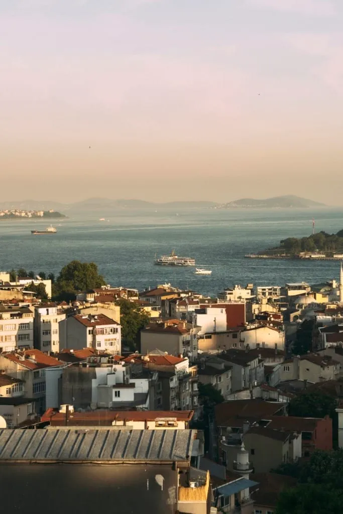 Bir şehir silüetini ve geniş bir su yolunu gösteren bu dikey çekimde, ön planda güneş ışığıyla aydınlanmış, bitişik nizamda sıralanmış çok katlı ve kiremit çatılı binalar yer alıyor; ortadaki masmavi denizde bir vapur ve küçük tekneler hareket ederken, uzakta hafif puslu tepeler ve adalar seçiliyor.