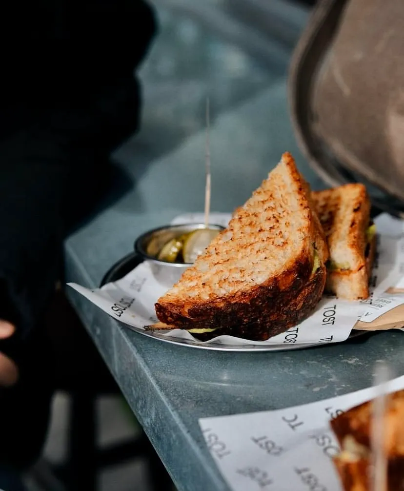 Koyu renkli bir masanın üzerinde, metal bir tabakta TOST yazılı kağıtla sunulan, mükemmel şekilde kızarmış, altın rengi üçgen organik tost dilimleri ve yanında turşu dolu küçük bir kaseyle iştah açıcı bir sunum.