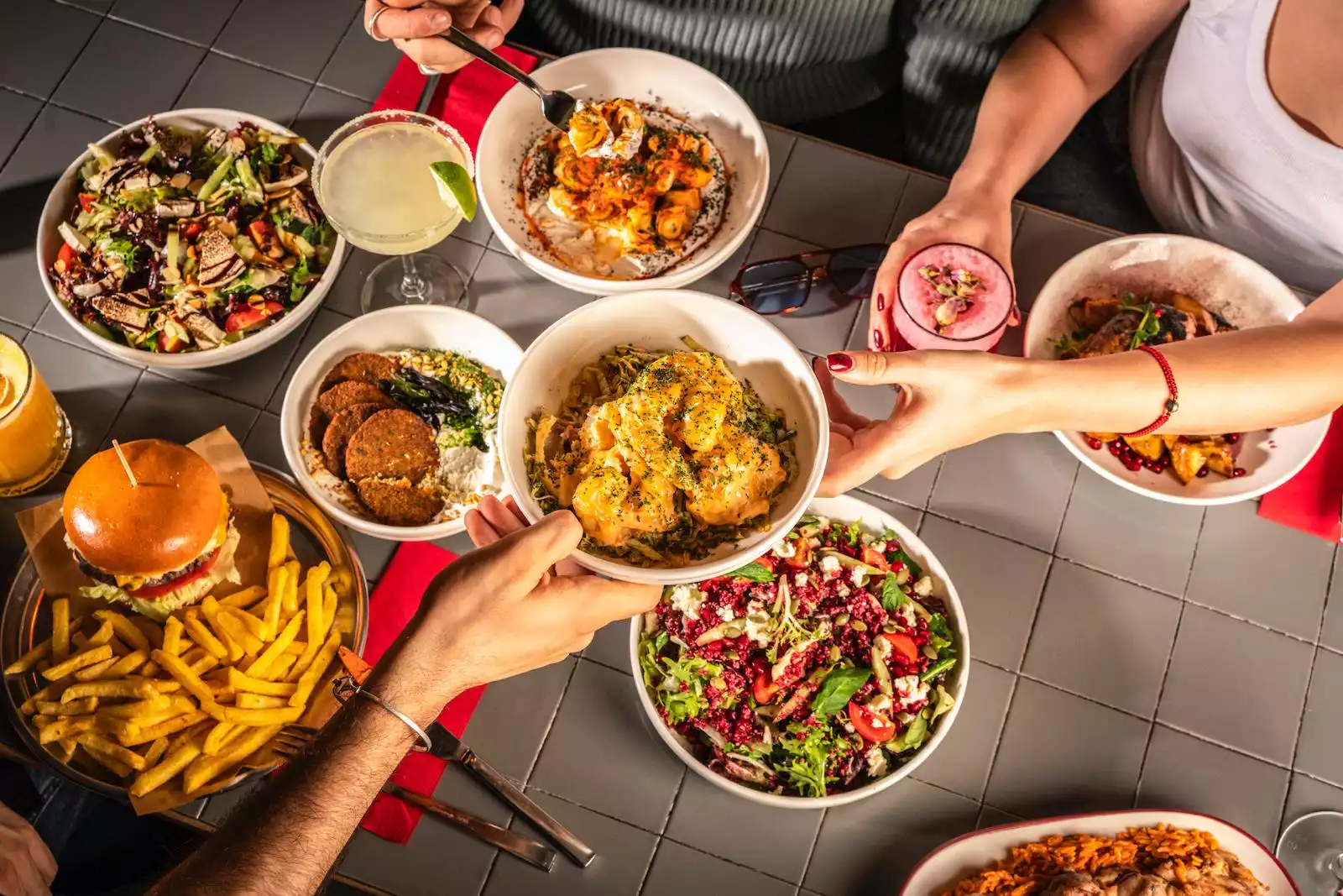 Oggusto - İstanbul’un En Lezzetli Street Food Restoranları: Gri fayanslı masada buluşan dostlar; dev hamburger, patates kızartması, falafel ve taze salataları paylaşıyor. İştah açıcı bir kaseyi elden ele uzatarak şehrin dinamik ve paylaşıma dayalı samimi sokak lezzeti kültürünü yansıtıyorlar.