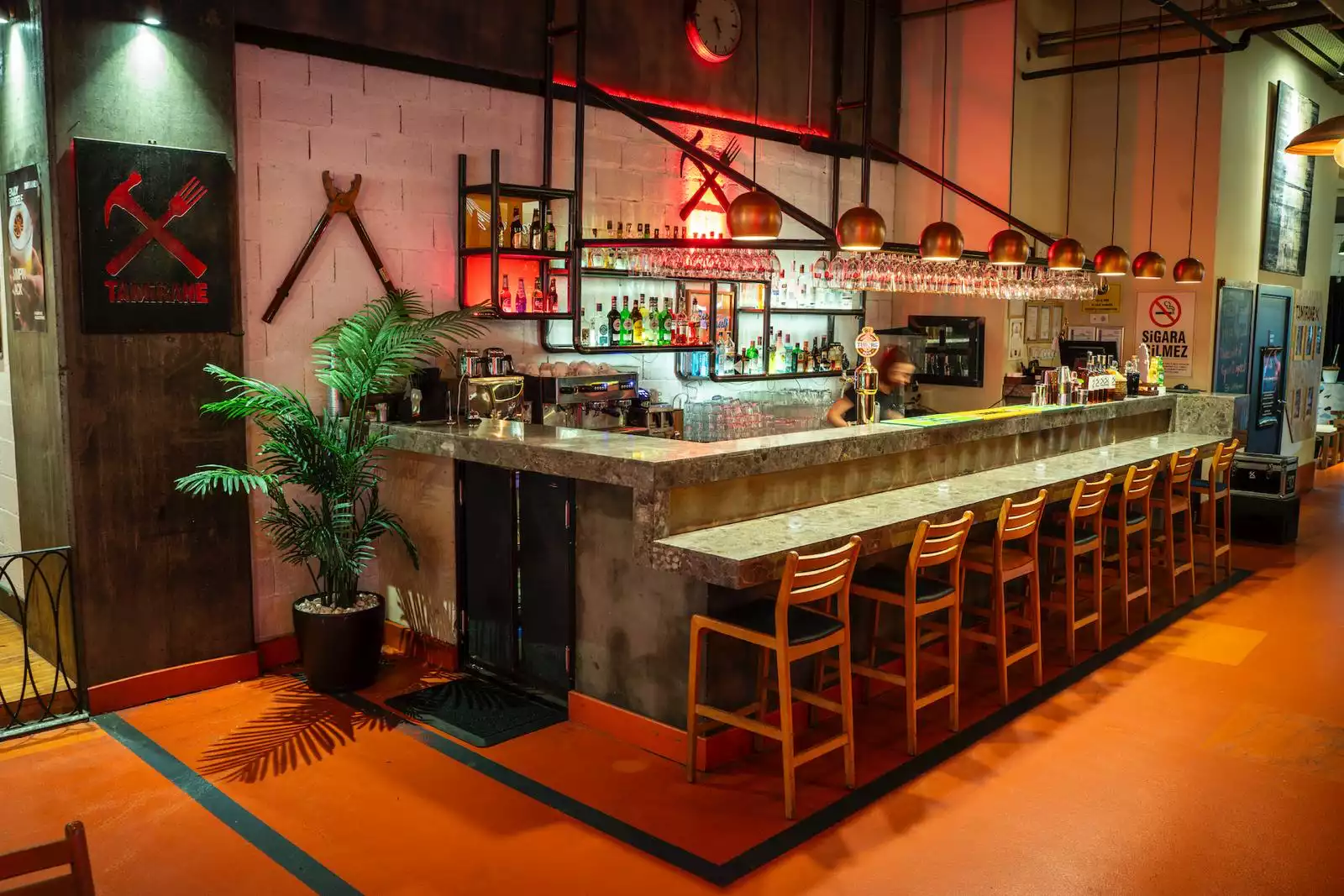 Oggusto - İstanbul’un En Lezzetli Street Food Restoranları: Tamirane mekanının endüstriyel bar alanı. Çapraz çekiç-çatal logolu duvar, uzun mermer tezgah, ahşap tabureler, bakır sarkıt lambalar ve kırmızı ışıklar, şehrin modern sokak lezzeti dinamizmini ziyaretçilere yansıtıyor.