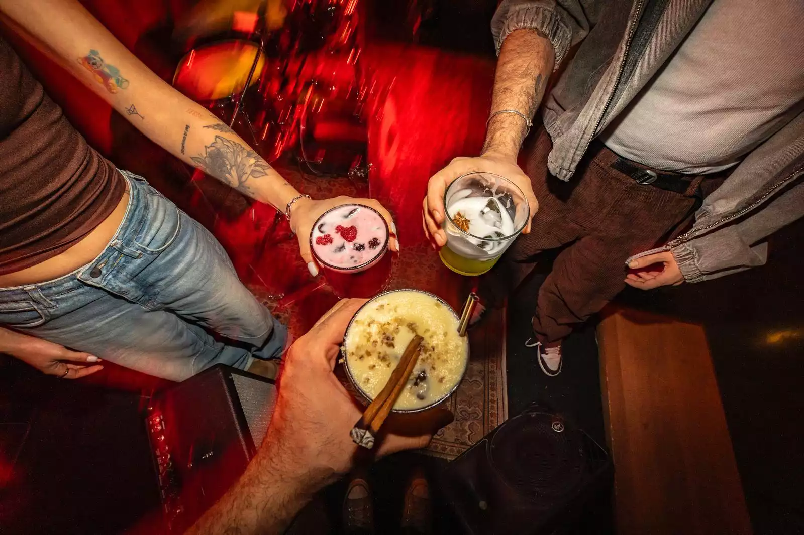 Oggusto - İstanbul’un En Lezzetli Street Food Restoranları rehberinden: Kırmızı loş ışıklı ve enerjik bir atmosferde, sokak lezzetlerine eşlik eden butik kokteyllerle kadeh kaldıran üç genç. İçeceklerin üzerindeki kırmızı meyveler, yıldız anason ve tarçın çubuğu sunuma zenginlik katıyor.
