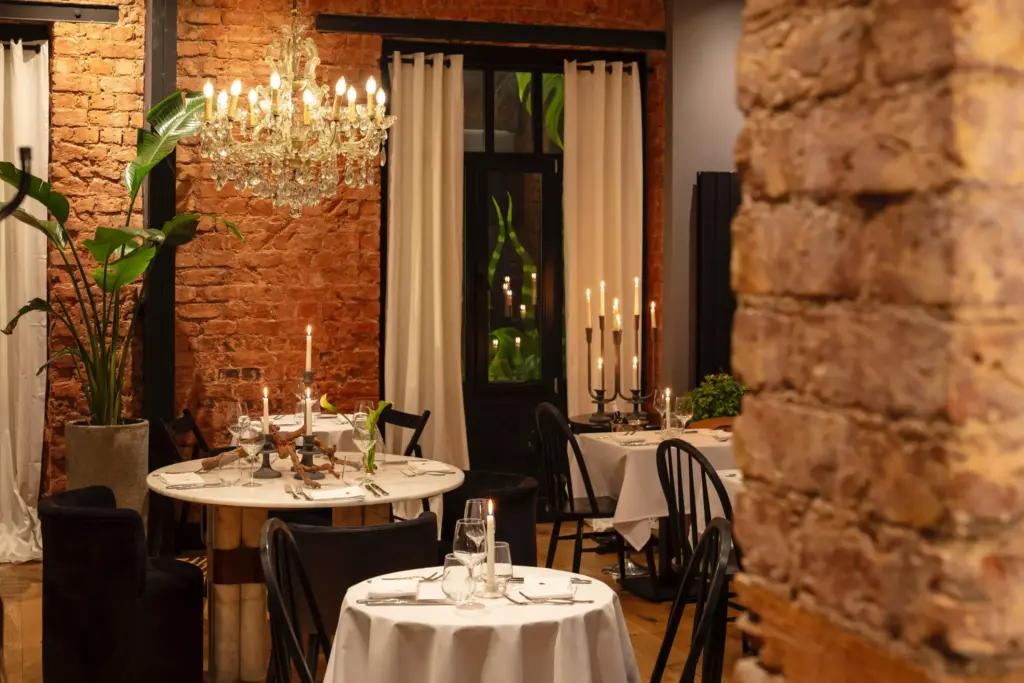 Tuğla duvarlı, kristal avizelerle aydınlatılmış şık bir restoran veya bar iç mekanında, beyaz örtülü masalar özenle hazırlanmış, her birinde yanan mumlarla donatılmış şamdanlar ve yemek takımları yer alırken, arka planda beyaz perdelerle çevrili bir kapı ve büyük bir saksı bitkisi dikkat çekiyor, samimi ve davetkar bir atmosfer sunuyor.