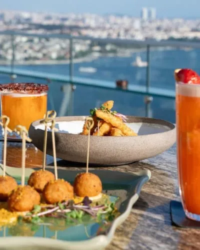 Rooftop restoran manzaralı masa ve kokteyller – İstanbul’un tarihi binaları manzarasına karşı servis edilen şık tabaklar ve kokteyl sunumu.