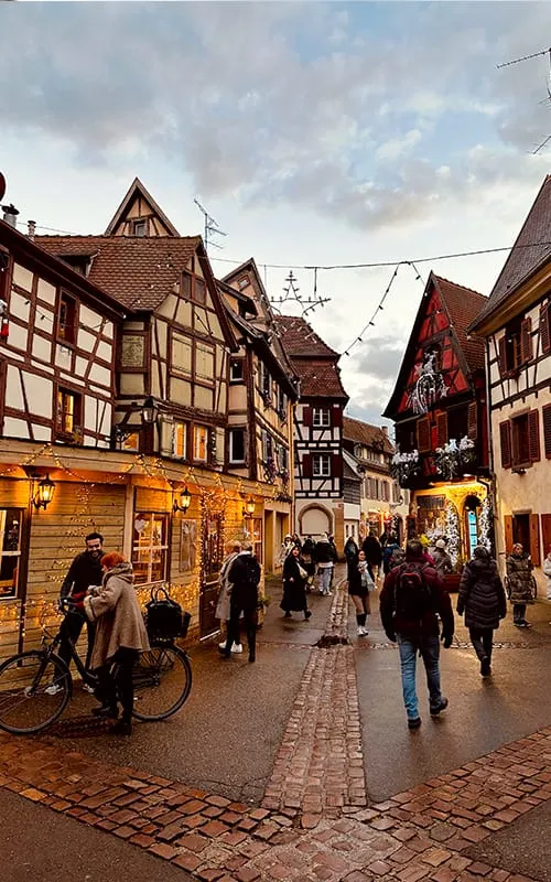 Colmar’da Alsace Noel Pazarları döneminde ışıklarla süslenmiş tarihi yarı ahşap evler ve sokakta gezen insanlar.