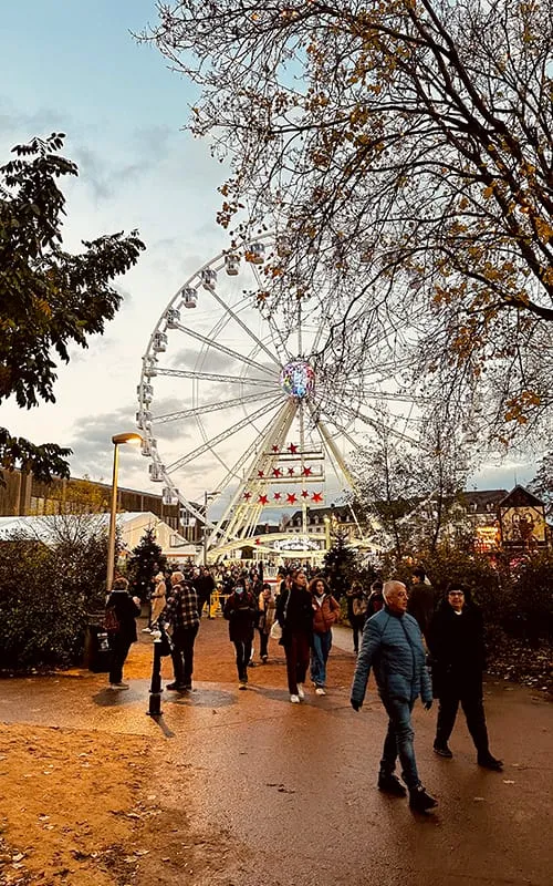 Alsace Noel Pazarları’nda yer alan büyük dönme dolap, yılbaşı ışıkları ve kalabalık festival alanında yürüyen insanlar.