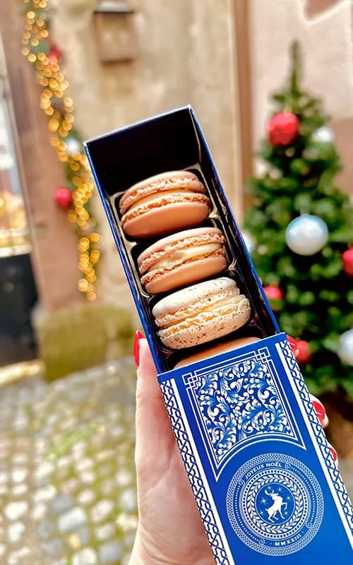 Alsace yılbaşı pazarlarında satılan geleneksel macaronların kutuda sunumu; arka planda Colmar’ın yılbaşı süslemeli taş sokakları ve Noel ağacı.