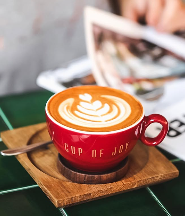 Bulanık arka planda bir dergiye göz gezdiren kişinin önünde, Cup of Joy kafesine özgü, üzerinde kalp şeklinde latte art deseni ve kırmızı fincanında CUP OF JOY yazısı bulunan, ahşap altlığı ve kaşığıyla birlikte koyu yeşil fayanslı bir masada duran sıcak bir kahve, Bebek'in keyifli mekanlarından birinde dinlendirici bir anı yansıtıyor.