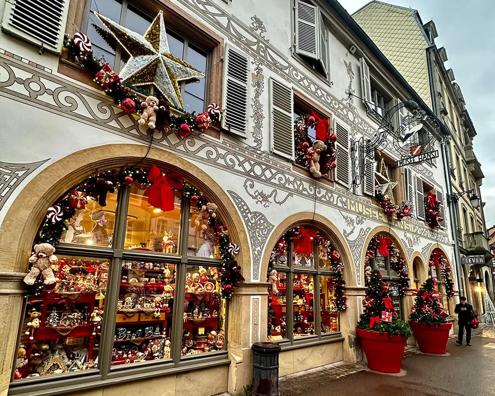 Colmar’da Musée Hansi’nin yılbaşı süslemeleriyle dekore edilmiş cephesi, büyük kırmızı saksılar, ışıklı çelenkler ve Noel temalı vitrinlerle Alsace bölgesinin Noel pazarı atmosferi.