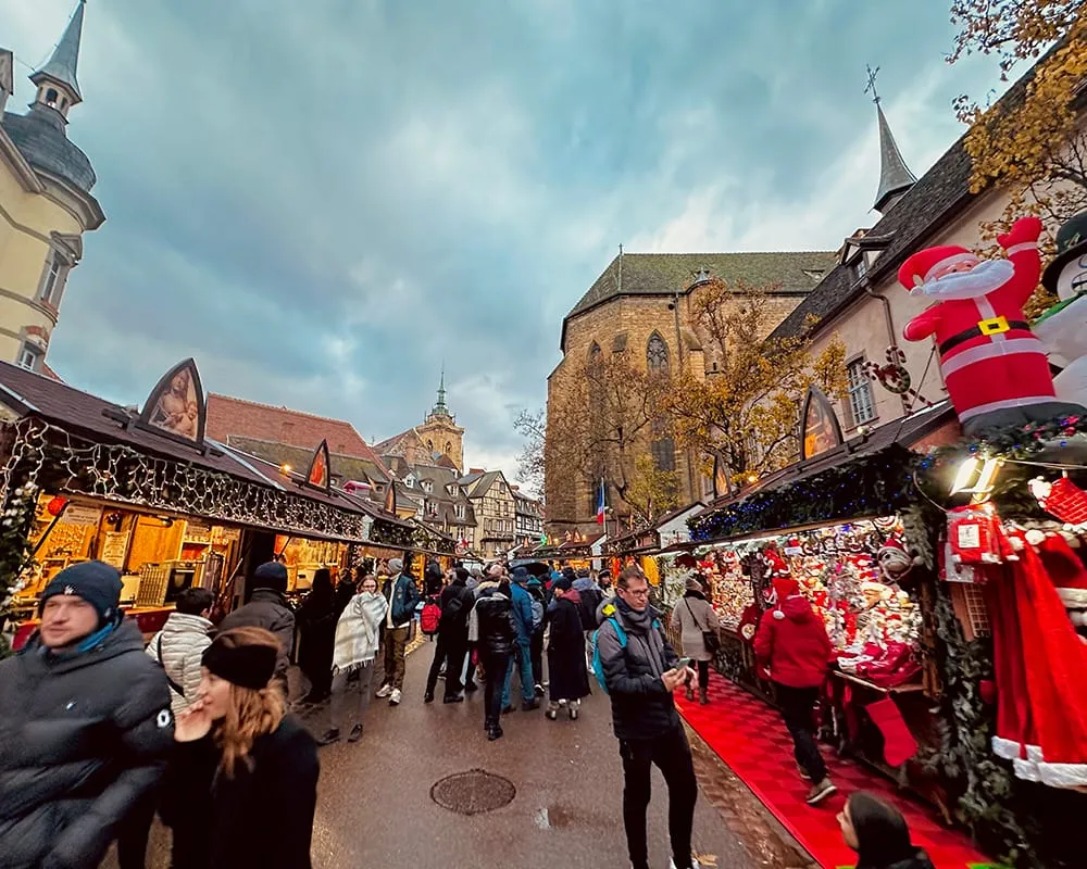 Colmar’da Alsace Noel Pazarları sırasında kırmızı süslemeli tezgâhlar, kalabalık ziyaretçiler ve katedralin gölgesinde kurulan Christmas Market atmosferi.