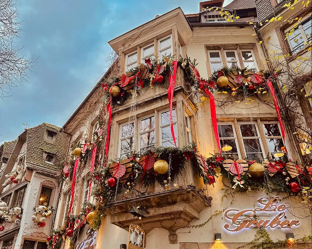 Colmar’da Alsace Noel Pazarları döneminde yoğun kırmızı kurdelelerle, çam dallarıyla ve altın süslerle dekore edilmiş tarihi Alsace evleri ve yılbaşı atmosferi.