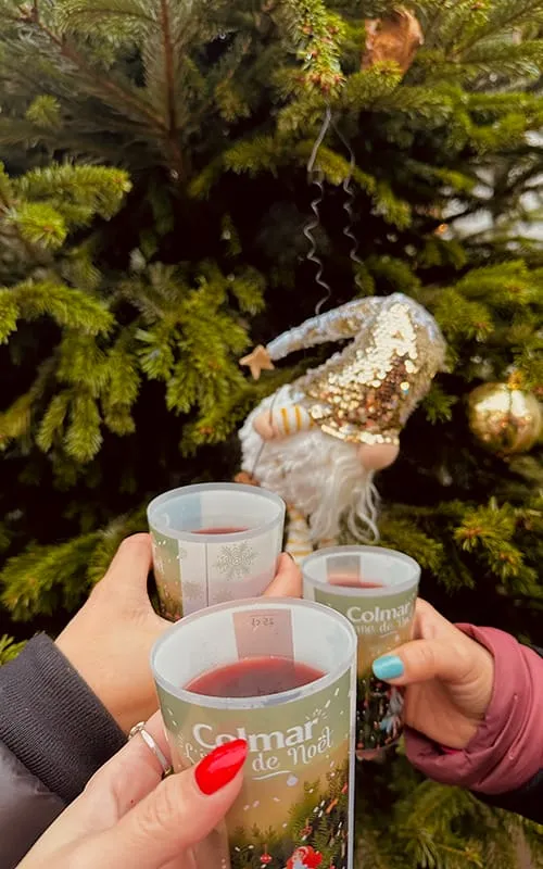 Colmar’da Alsace Noel Pazarları sırasında yılbaşı ağacının önünde üç kişinin vin chaud (sıcak şarap) bardaklarıyla kutlama yaptığı Christmas atmosferi.