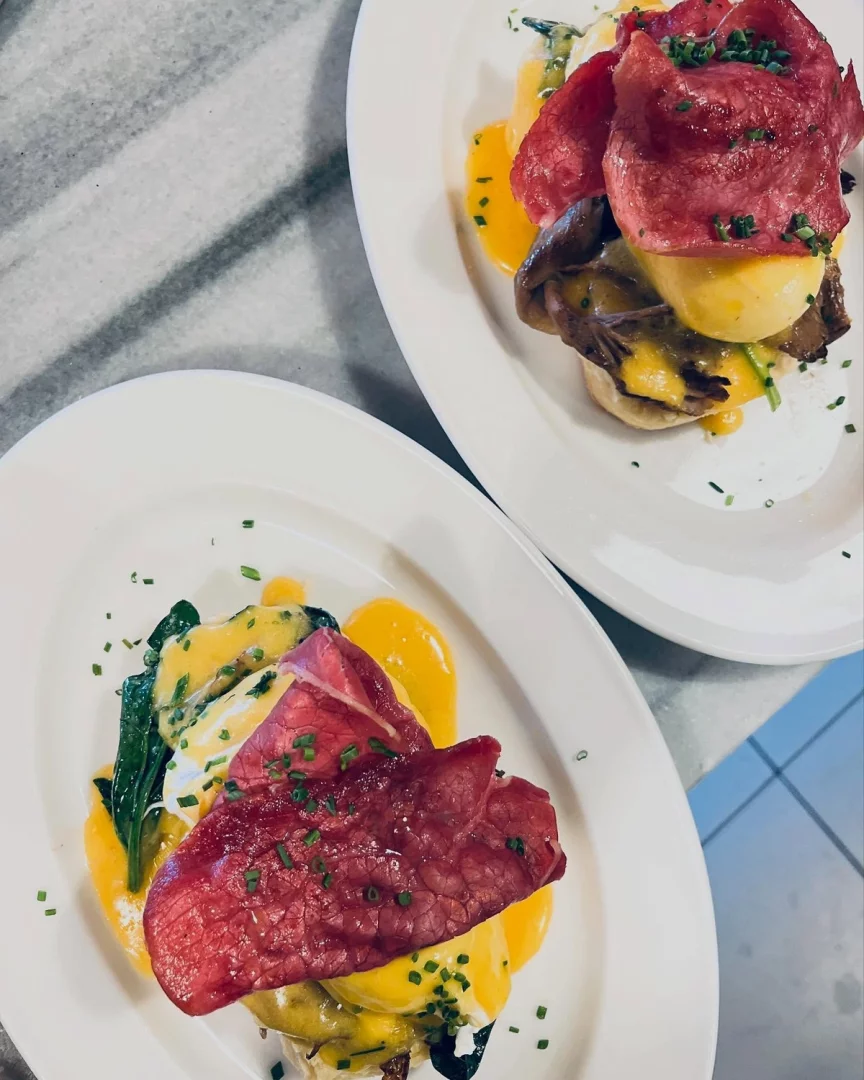 Cihangir'deki Cuppa'dan iki porsiyon Eggs Benedict, mermer yüzeyde üstten çekilmiş bir karede, poşe yumurta, bol Hollandez sosu, kırmızı pastırma dilimleri ve taze frenk soğanıyla bezenmiş, biri ıspanaklı diğeri mantarlı gibi görünen iştah açıcı kahvaltı tabaklarını gösteriyor.
