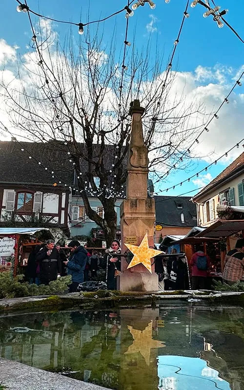 Eguisheim’da Alsace Noel Pazarları döneminde meydandaki tarihi çeşme, yıldız şeklinde yılbaşı süsleri, stantlar ve kalabalık Christmas Market atmosferi.