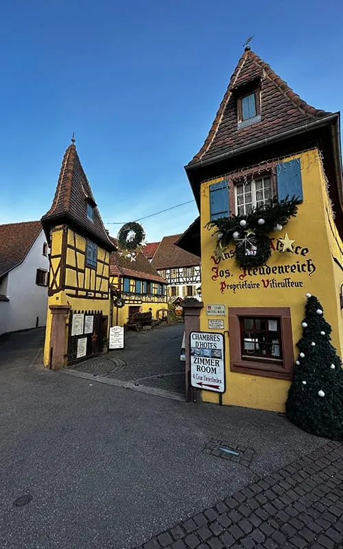 Eguisheim’da Alsace Noel Pazarları döneminde görülen sarı yarı ahşap tarihi evler, giriş kapısı ve yılbaşı süslemeleriyle bezeli sokak görünümü.