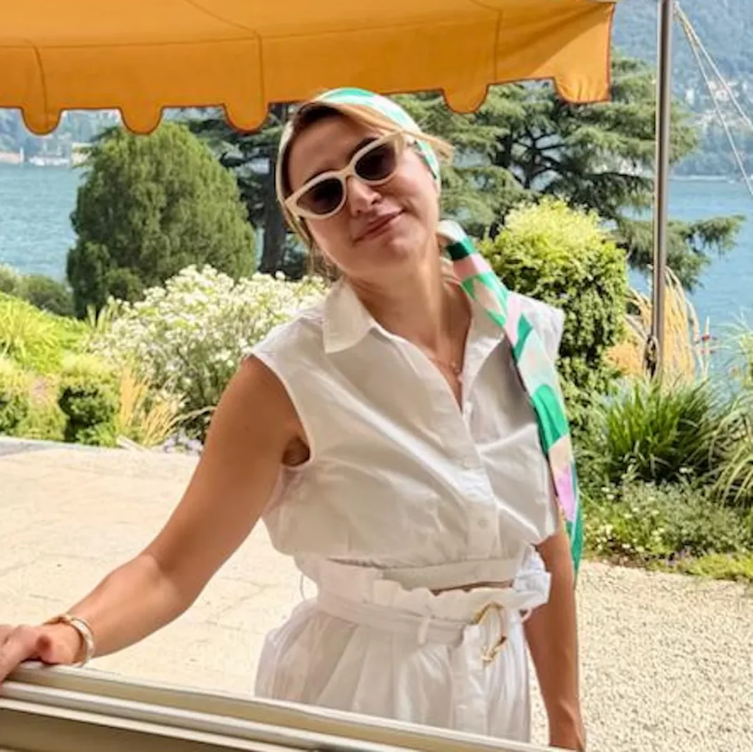 Fonksiyonel tıp sağlık koçu Ebru Zeynep Altay, güneşte parlayan göl manzarası ve yemyeşil doğa eşliğinde, beyaz kolsuz bluzu, pantolonu, kedi gözü güneş gözlükleri ve renkli eşarbıyla huzurlu bir ifadeyle gülümsüyor, sağlıklı yaşam felsefesini zarifçe yansıtıyor.