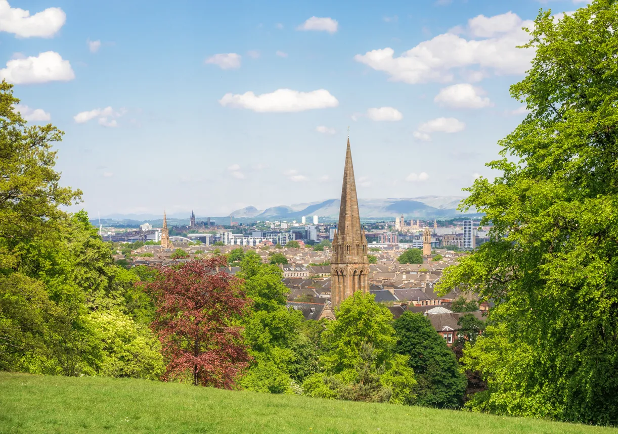 Glasgow şehrinin güneşli bir günde çekilmiş bu geniş manzarasında, ön planda yemyeşil çimenler ve farklı tonlarda ağaçlar, orta kısımda kentin üzerinden Gotik tarzı sivri bir taş kilise kulesi ve diğer binalar yükselirken, ufukta dağ silüetleri mavi gökyüzü ve beyaz bulutlarla birleşiyor.