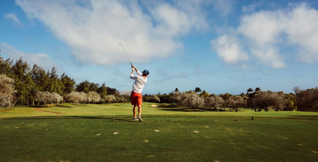 Güneşli bir günde yemyeşil bir golf sahasında, beyaz tişörtlü, kırmızı şortlu ve mavi şapkalı bir oyuncu, golf sopasını kuvvetlice savurarak vuruşunu tamamlıyor, arka planda ise gür ağaçlar ve hafif bulutlu mavi bir gökyüzü uzanıyor.