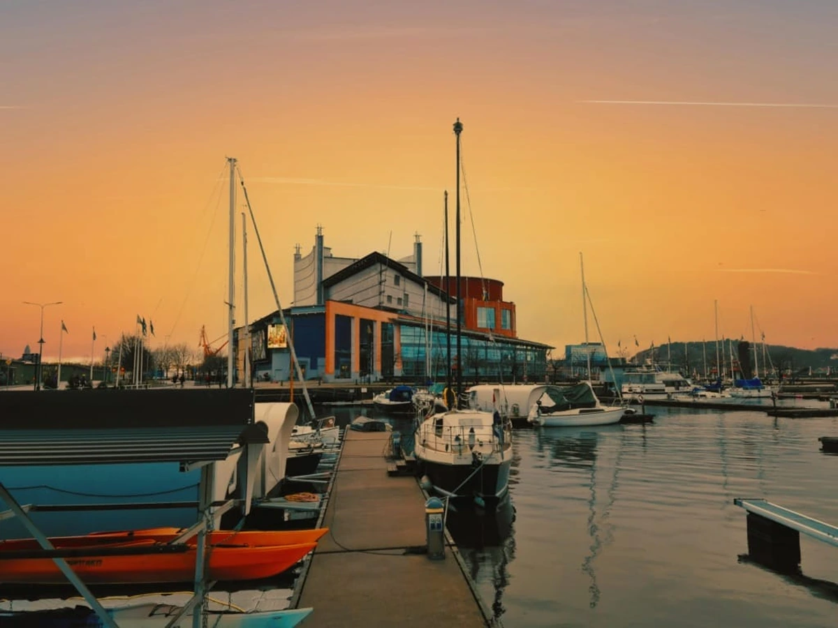 Gün batımının turuncu ve mor tonlarının yansıdığı sakin bir Göteborg marinasında, modern bir binanın önünde demirli çok sayıda yelkenli ve küçük tekneyle, sürdürülebilir seyahat rotasına vurgu yapan huzurlu bir liman manzarası.