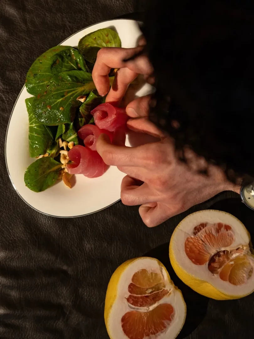 Cihangir'deki Hona restoranının özgün sunumlarından biri olarak, bir elin koyu renkli bir masanın üzerindeki beyaz tabağa parlak yeşil yapraklı salata ve fıstıkların arasına özenle pembe, rulo şeklinde dilimlenmiş ton balığı parçalarını yerleştirdiği anı gösteren, yanında ikiye bölünmüş pembe içli pomelo dilimleri olan dikkat çekici bir yemeğin görüntüsü.