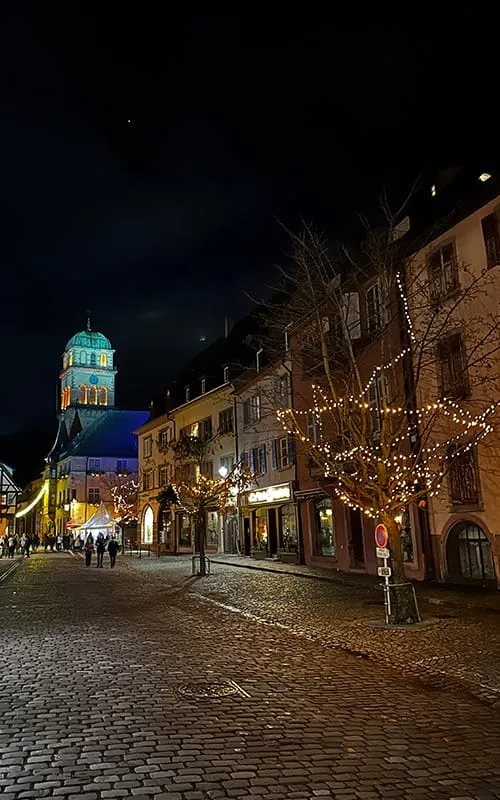 Kaysersberg’de Alsace Noel Pazarları döneminde gece ışıklarıyla süslenen taş sokaklar, aydınlatılmış katedral ve yürüyen ziyaretçiler.