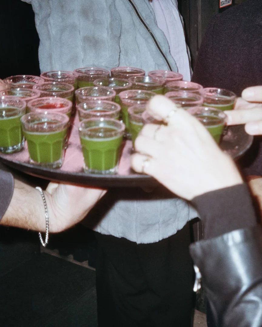 Servis görevlisinin tuttuğu metal bir tepside, çoğu canlı yeşil renkteki enerji verici matcha shotları ve aralarına karışmış birkaç kırmızı shot, yüzüklü bir el bir içeceğe uzanırken anlık bir karede belirginleşiyor; bu canlı sunum, loş ışıklı bir etkinlik ortamını çağrıştırıyor.
