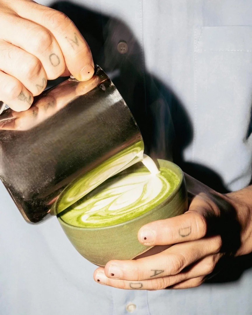 Ellerinde dikkat çekici dövmeler bulunan bir barista, Le Oba kahve evinin yeşil seramik kasesindeki parlak yeşil matcha latte üzerine metal bir sürahiden buharla ısıtılmış sütü hassasiyetle dökerek zarif yaprak deseni şeklinde latte sanatı oluşturuyor.