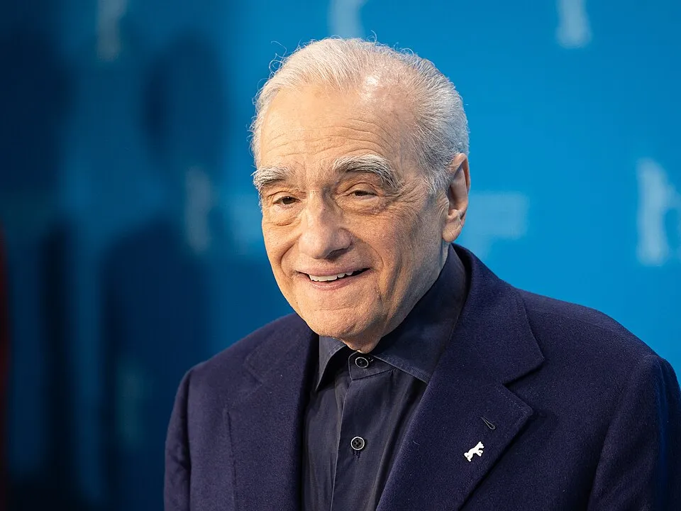 Sinema dünyasının önemli isimlerinden yönetmen Martin Scorsese, lacivert ceketi ve yakasındaki Berlinale ayısı rozetiyle, mavi tonlarda bulanık bir etkinlik arka planı önünde, sıcak bir gülümsemeyle poz veriyor.