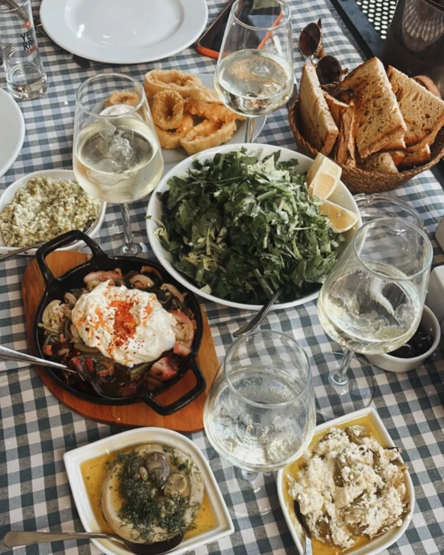 Mavi beyaz kareli bir masa örtüsü üzerinde, bir meyhane ortamında servis edilen taptaze roka salatası, çıtır kalamar tava, enginar, yoğurtlu sebze ve ekmek gibi çeşitli mezelerle donatılmış masa, beyaz şarap kadehleriyle doludur ve zengin bir ziyafeti gözler önüne serer.