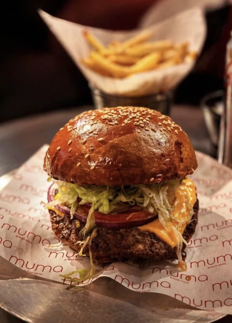 Yakın çekim bir fotoğraf, susamlı parlak bir burger ekmeği arasında kıvırcık marul, kırmızı soğan, domates dilimleri, erimiş sarı peynir ve bol etli bir köftenin yer aldığı Muma Burger House'a ait lezzetli bir burgeri gösteriyor; arka planda bulanık bir şekilde patates kızartmaları seçiliyor.