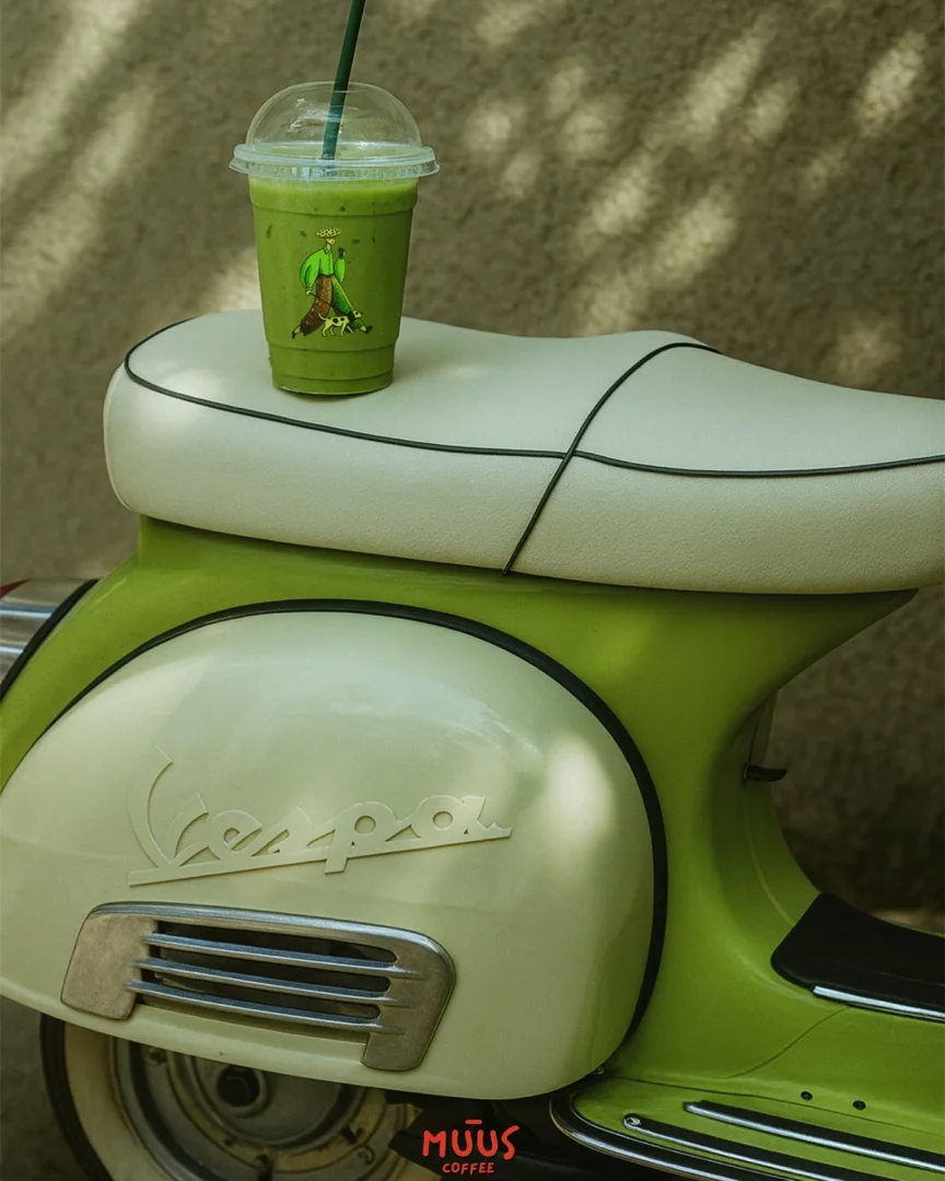 Canlı yeşil rengiyle dikkat çeken, üzerinde karakter çizimi bulunan bir Muus Coffee matcha içeceği, krem ve yeşil tonlarındaki şık bir Vespa motosikletin bej renkli sele koltuğunun üzerinde duruyor; bu görüntü, Vespa ve matcha keyfini bir araya getiriyor.
