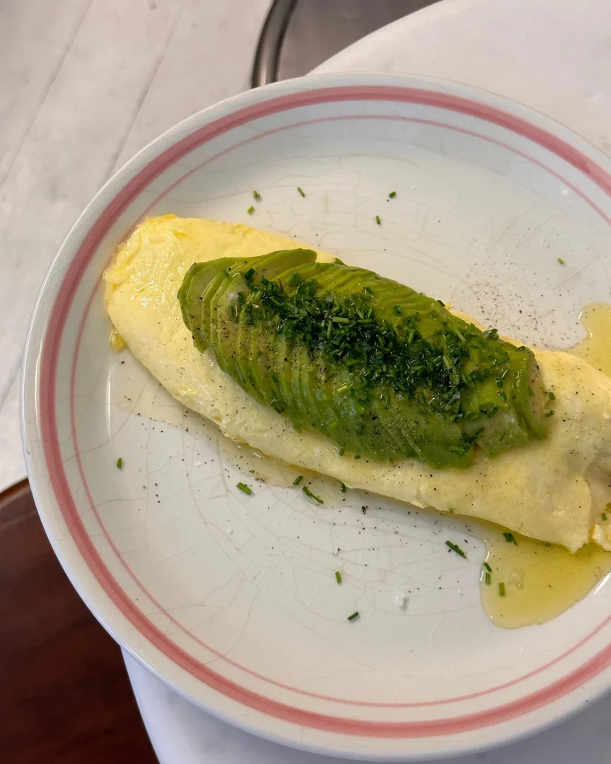 Beyaz, pembe kenarlı ve çatlak dokulu bir tabağın ortasında, üzerine ince dilimlenmiş parlak yeşil avokado, doğranmış frenk soğanı ve taze çekilmiş karabiber serpilmiş, kenarlarından hafifçe yağ sızan iştah açıcı bir avokadolu omlet.