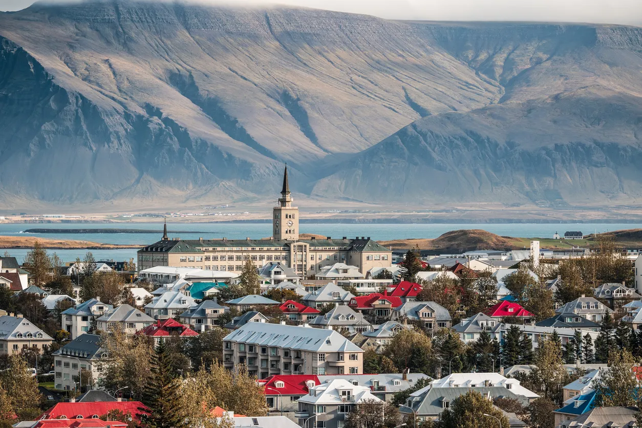 Reykjavik şehrinin, ön planda renkli çatılı evleri, orta planda saat kulesiyle öne çıkan üniversite binası ve arka planda masmavi deniz ile yükselen heybetli dağlarıyla panoramik bir görünümü.