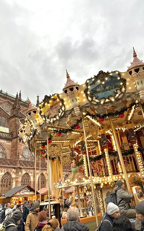 Strasbourg’da Alsace Noel Pazarları döneminde Notre Dame Katedrali önünde kurulan ışıklı atlı karınca (carousel) ve kalabalık yılbaşı pazarı atmosferi.