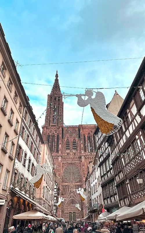 Strasbourg’da Alsace Noel Pazarları döneminde Notre Dame Katedrali’ne uzanan caddede asılı melek süslemeleri, kalabalık ziyaretçiler ve tarihi Alsace evleri.