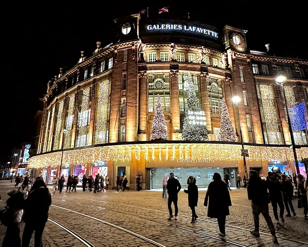 Strasbourg’daki Galeries Lafayette binasının yılbaşı ışıkları ve dev Noel ağaçlarıyla süslenmiş gece görüntüsü; Alsace Noel pazarları döneminin şehir atmosferi.