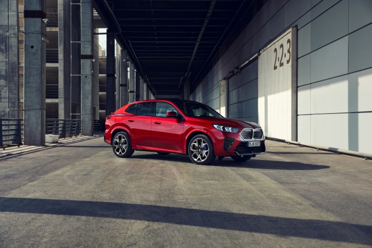 Kırmızı BMW iX2 elektrikli SUV, modern endüstriyel mimari önünde şehir atmosferinde park halinde.