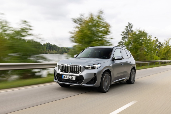 Gri BMW iX1 elektrikli SUV, ağaçlarla çevrili yolda sürüş halinde dinamik tasarımıyla öne çıkıyor.