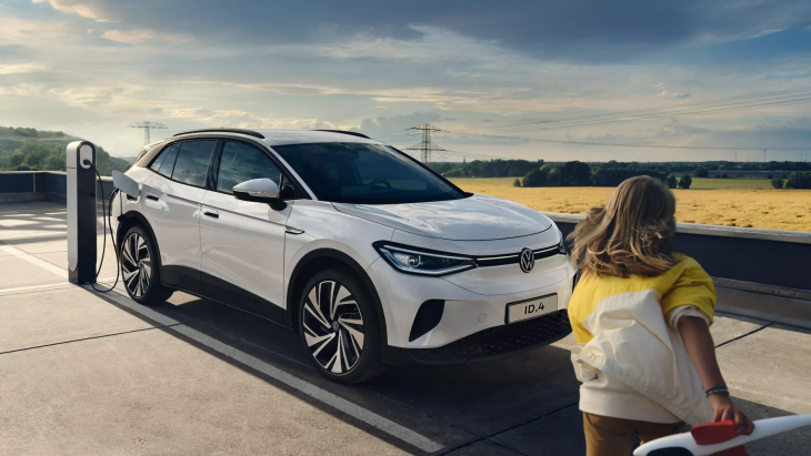 Şarj istasyonuna bağlı beyaz Volkswagen ID.4 elektrikli SUV, açık alanda park halinde; yanında çocuklu bir aile detayı.