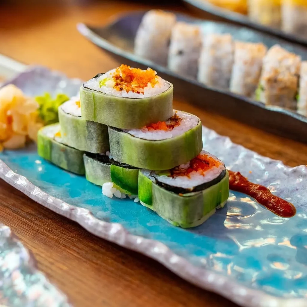 İstanbul'un en iyi sushi restoranları arasında öne çıkan, turkuaz bir tabakta sunulan, dışı taze salatalık dilimleriyle sarılı, içi pirinç ve nori ile dolu, üzeri canlı turuncu balık yumurtası ve baharatlı sosla zenginleştirilmiş, estetik ve inovatif bir yaklaşımla yığılmış lezzetli sushi roll'ları.