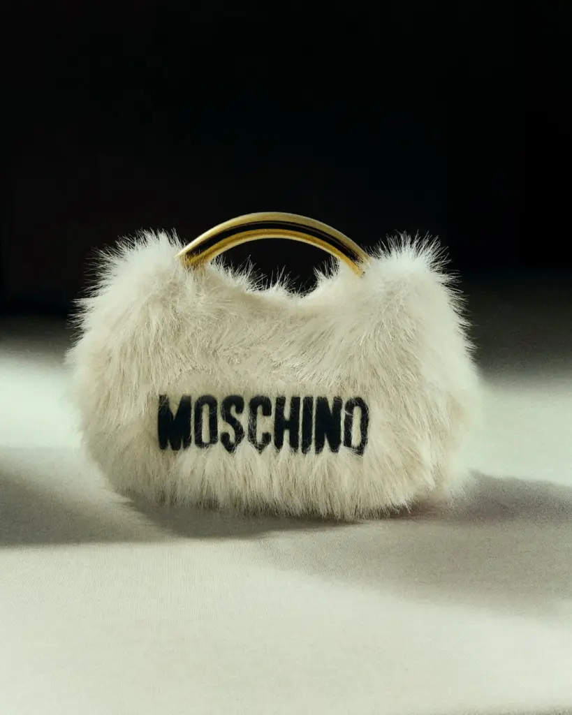 Beyaz yapay kürkle kaplı, altın saplı Moschino el çantası stüdyo ortamında aydınlık bir zeminde sergileniyor.