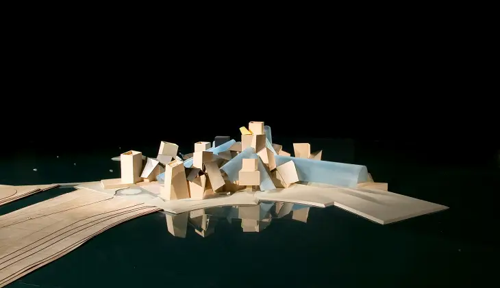 Karanlık ve yansıtıcı bir zemin üzerinde, Frank Gehry'nin imzasını taşıyan, Guggenheim Abu Dhabi Müzesi'nin farklı yükseklik ve şekillerdeki galerilerini temsil eden, birbiriyle iç içe geçmiş kümelenmiş kutu benzeri yapı elemanlarından oluşan ve bazı bölümlerinde şeffaf mavi detaylar barındıran soyut bir mimari maket.