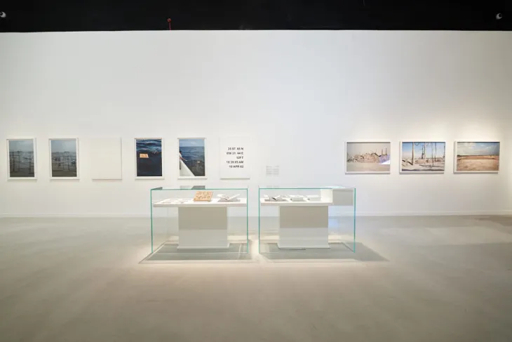 Guggenheim Abu Dhabi Müzesi'nin geniş ve aydınlık bir sergi salonunda, modern sanat eserleri olarak duvarlarda deniz, kentsel ve çöl görünümleri ile coğrafi koordinatlar içeren fotoğraflar asılı; zemindeki cam vitrinlerde ise tamamlayıcı belgeler ve küçük ölçekli objeler sergileniyor.