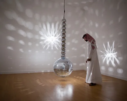 Guggenheim Abu Dhabi Müzesi'nin ışık temalı bir çağdaş sanat sergisinden, beyaz duvarlı ve ahşap zeminli bir galeride, geleneksel Arap kıyafetleri giymiş bir adamın tavandan sarkan, delikli küresel bir sanat eserinden yayılan ışık desenlerini incelediği büyüleyici bir an.