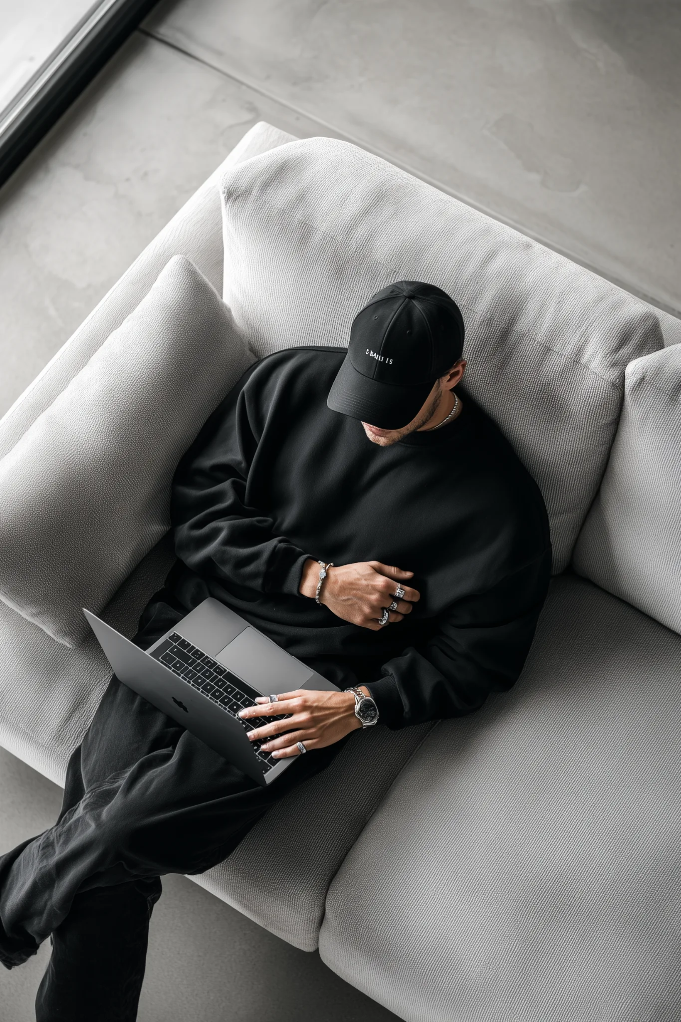 Minimalist salonda kanepede dizüstü bilgisayar kullanan erkek; siyah sweatshirt, şapka ve modern ev-ofis düzeni.