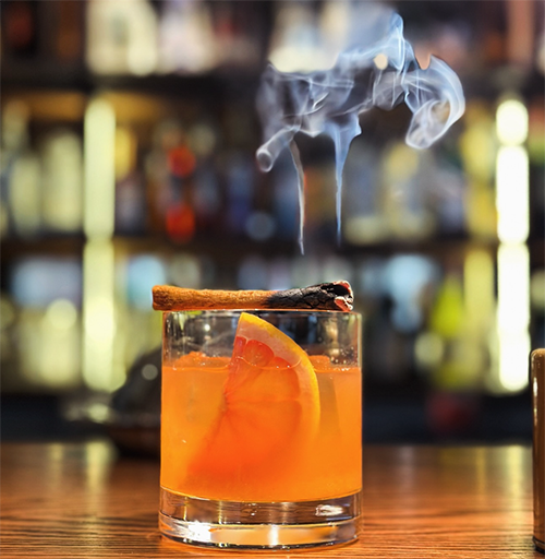 Bulanık ışıklandırılmış bir bar arka planında, ahşap tezgahın üzerinde duran kısa, şeffaf bir bardakta, parlak turuncu renkte bir kokteyl, bardağın kenarına yerleştirilmiş, hafifçe tüten ve dumanı yukarı doğru süzülen bir tarçın çubuğu ve içeceğin içinde bir dilim portakal kabuğu görünen çekici bir içecek.