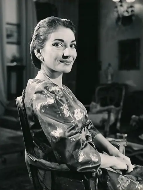 Maria Callas'ın hayatından bir kesit sunan bu siyah beyaz portrede, ünlü opera sanatçısı inci takıları ve zarif çiçek desenli saten bir elbiseyle süslü bir sandalyede oturmuş, hafifçe gülümseyerek objektife bakıyor.
