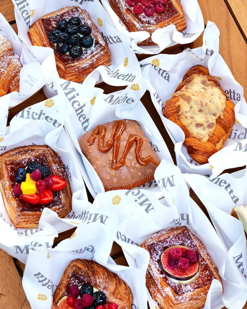 İstanbul'daki en iyi kruvasan duraklarından Michetta Bakery'nin beyaz logolu ambalajlarına sarılı, üzeri yaban mersini, karışık orman meyveleri, incir, ahududu ve çikolata gibi farklı dolgu ve süslemelerle bezeli, çıtır katmanlı çeşitli kruvasan ve hamur işlerinin iştah açıcı bir şekilde sergilendiği üst açı çekim.