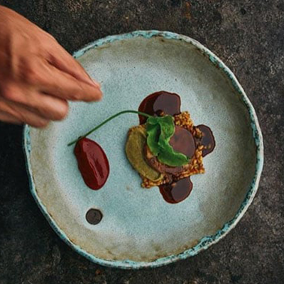 İstanbul'un Michelin Yıldızlı restoranlarından Neolokal'in özgün mutfak anlayışını yansıtan modern bir yemek sunumu: açık renk seramik tabakta, şefin titizlikle yeşil bir otu yerleştirdiği, zengin soslar, tahıllar ve koyu renkli ana bileşenlerle hazırlanmış, sanatsal ve estetik bir lezzet.