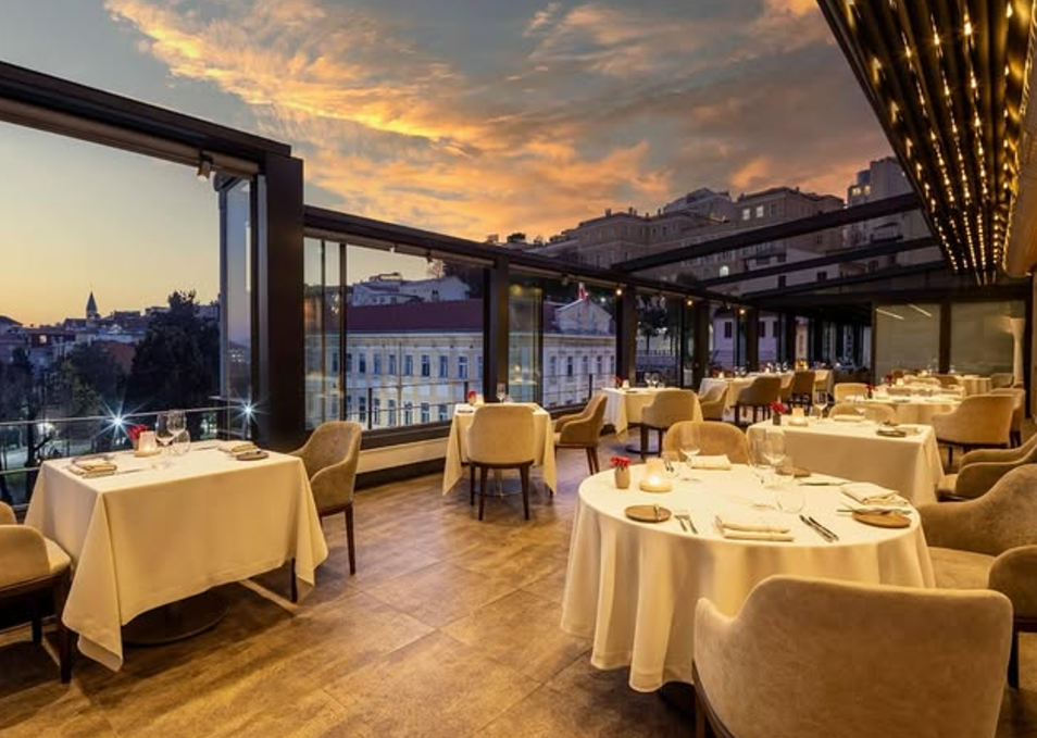 İstanbul'un Michelin Yıldızlı restoranlarından Nicole'ün gün batımı kızıllığıyla aydınlanan camlı terasında, şehir manzarasına karşı özenle düzenlenmiş masa ve sandalyelerle zarif bir yemek alanı.