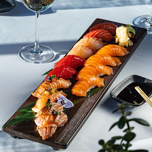 Narince hazırlanmış somon, orkinos ve közlenmiş balık nigiri çeşitlerinin yenilebilir çiçeklerle süslenerek sunulduğu uzun, koyu renk ahşap bir servis tabağı, İstanbul’un en popüler sushi restoranlarından birindeki zarif bir lezzet şölenini gözler önüne seriyor.